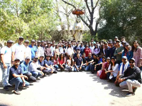 Large group photo of team members on a trip, gathered near a statue under trees
